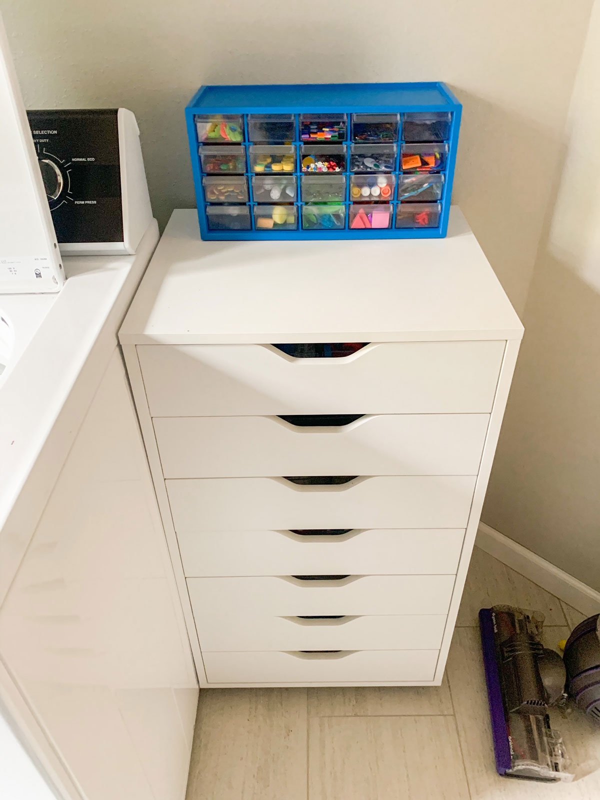 A white chest of drawers in a laundry room. On top is a blue set of drawers with craft supplies.