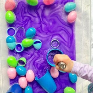 A child with a whisk is stirring purple metallic water in a sensory bin with Easter eggs.