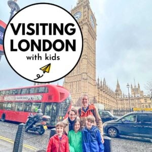 Visiting London with Kids: Images shows a family of 5 in front of Big Ben and the Elizabeth Tower. A double decker bus is passing behind the family.