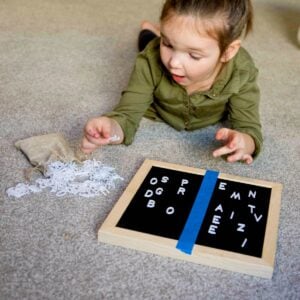A child laying on her stomach holds up a small white letter before placing it on a letter board. A blue piece of tape separates the board into two sides.