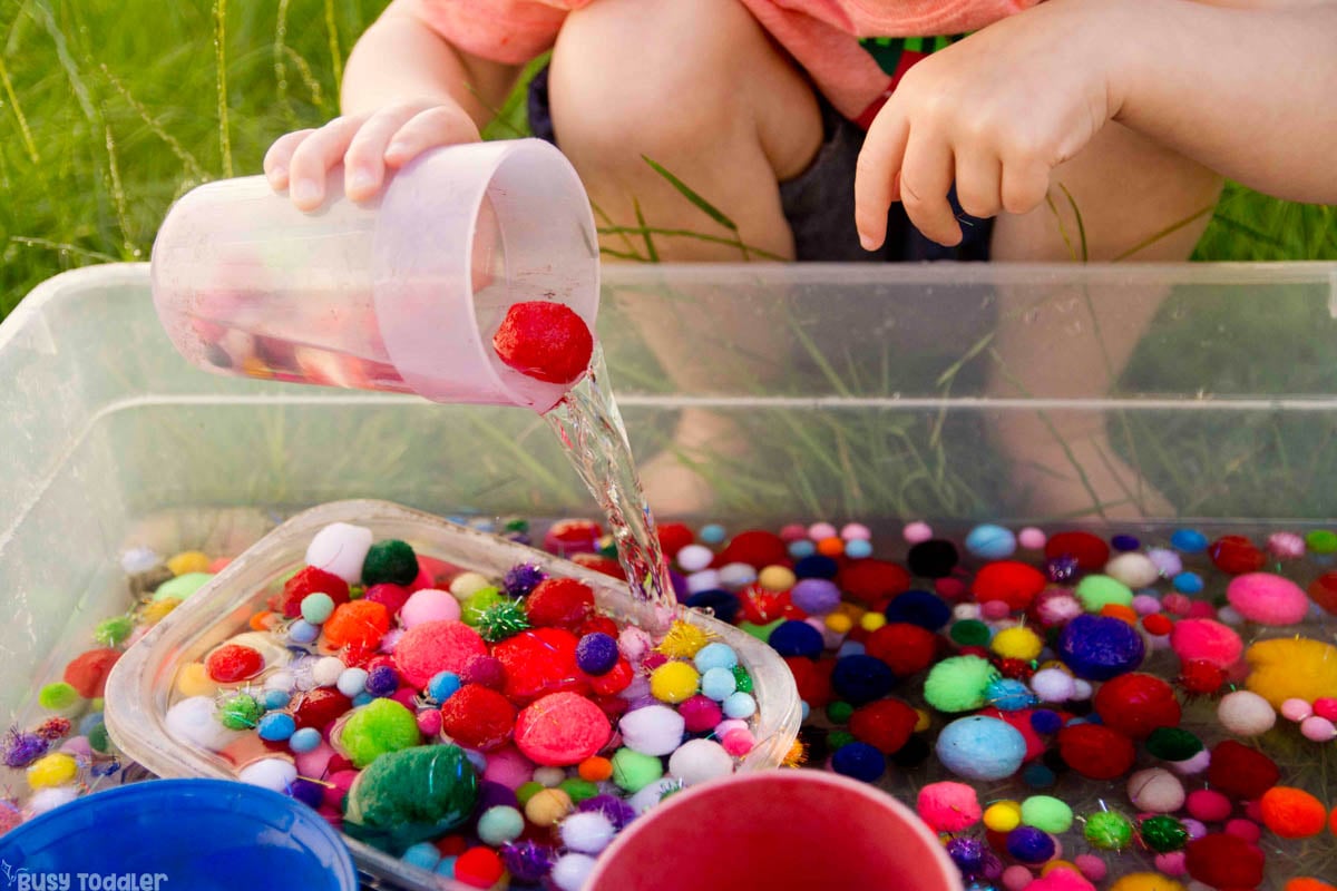 A child pours pom pom balls and water out of a cup into a sensory bin.