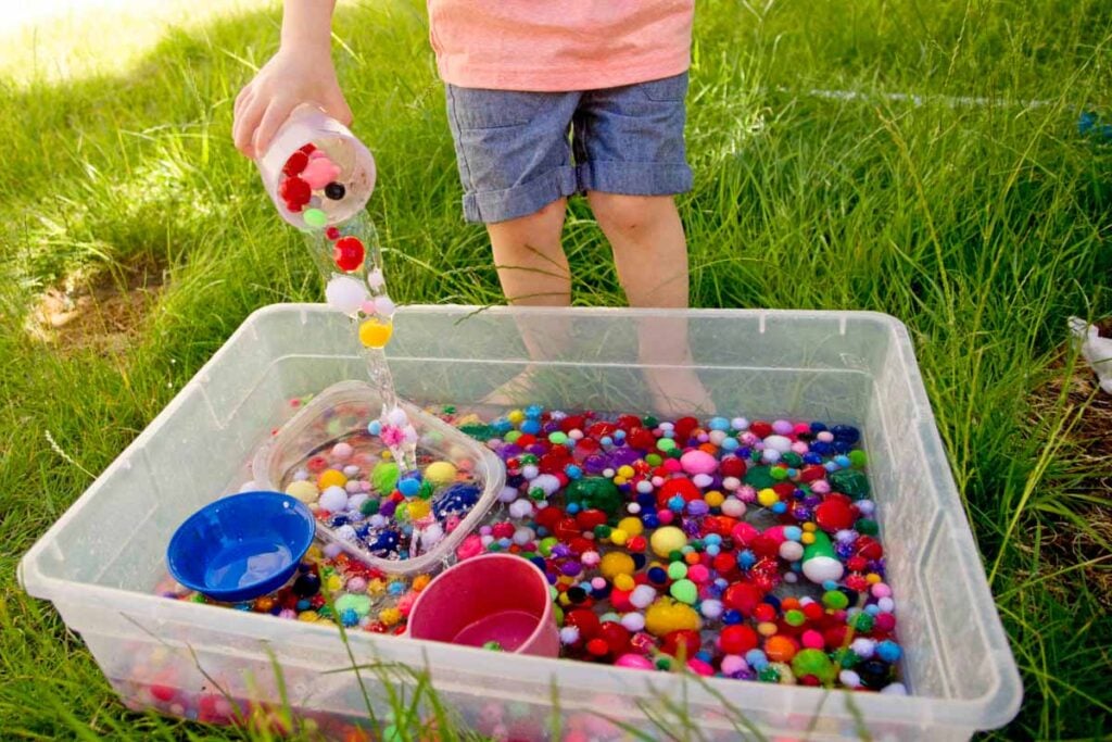 A child pours pom pom balls back into a sensory bin full of water.